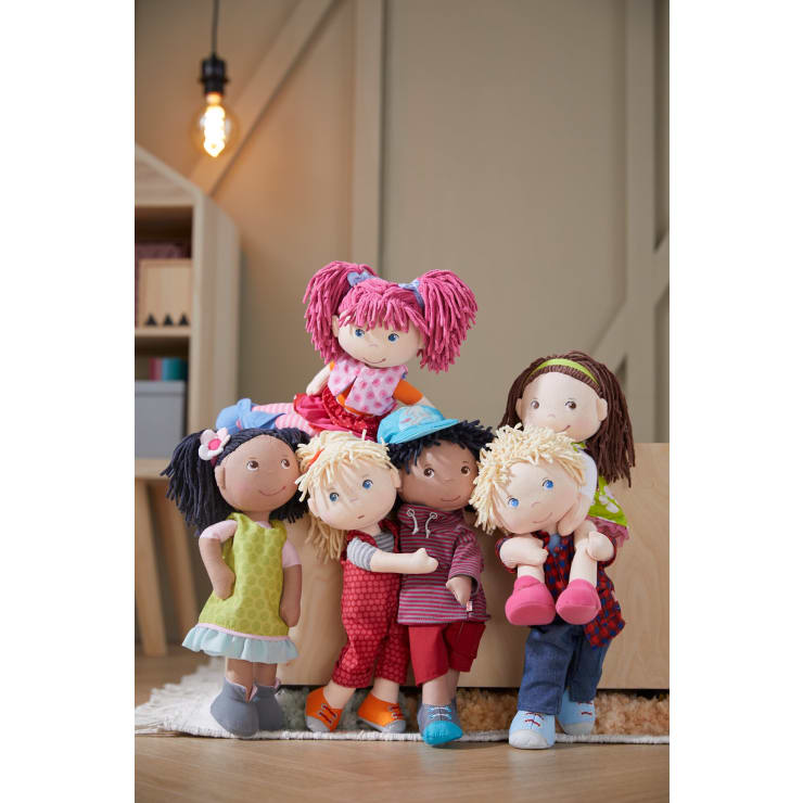 Sechs Stoffpuppen mit farbenfroher Kleidung und unterschiedlichen Haarfarben und Frisuren sind in einem Raum vor Holzmöbeln und einem Bücherregal unter einer beleuchteten Hängelampe gruppiert. Die Puppen sind in freundlichen Posen angeordnet.