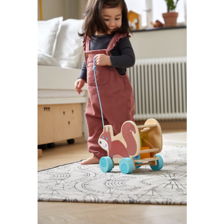 Ein kleines Kind in einem rosa Overall zieht ein Holzspielzeug in Form eines Eichhörnchens auf Rädern über einen gemusterten Teppich in einem hellen Zimmer mit einem Bett und einem Heizkörper im Hintergrund.