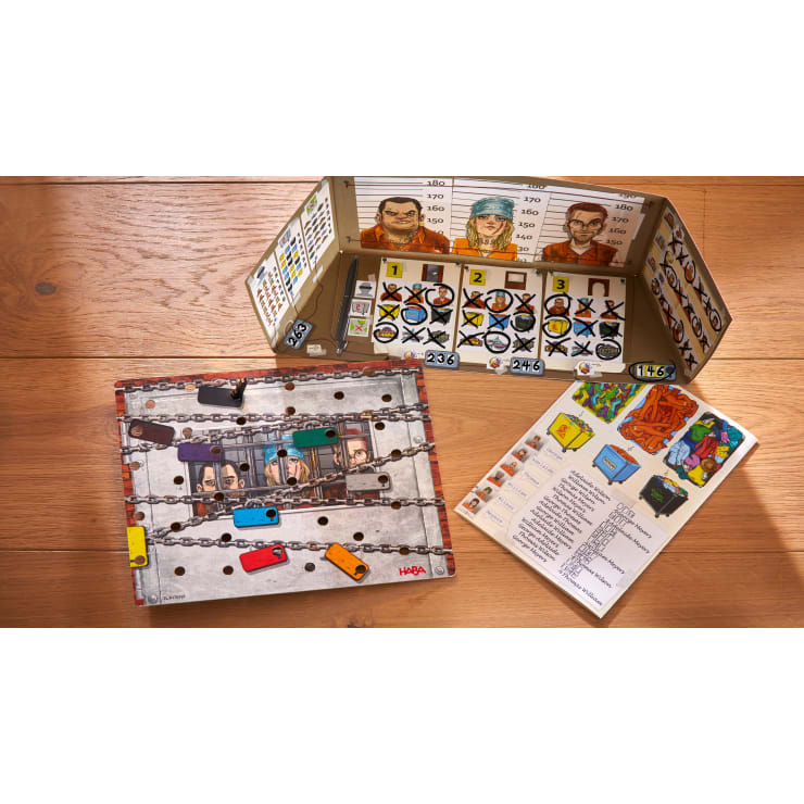 Ein Brettspiel mit einem Gefängnismotiv, das eine Anzeigetafel mit illustrierten Insassen, ein mit einem Vorhängeschloss versehenes Spielbrett, farbige Schlüssel und ein Anleitungsblatt mit Illustrationen und Anweisungen enthält, die alle auf einer Holzoberfläche angeordnet sind.