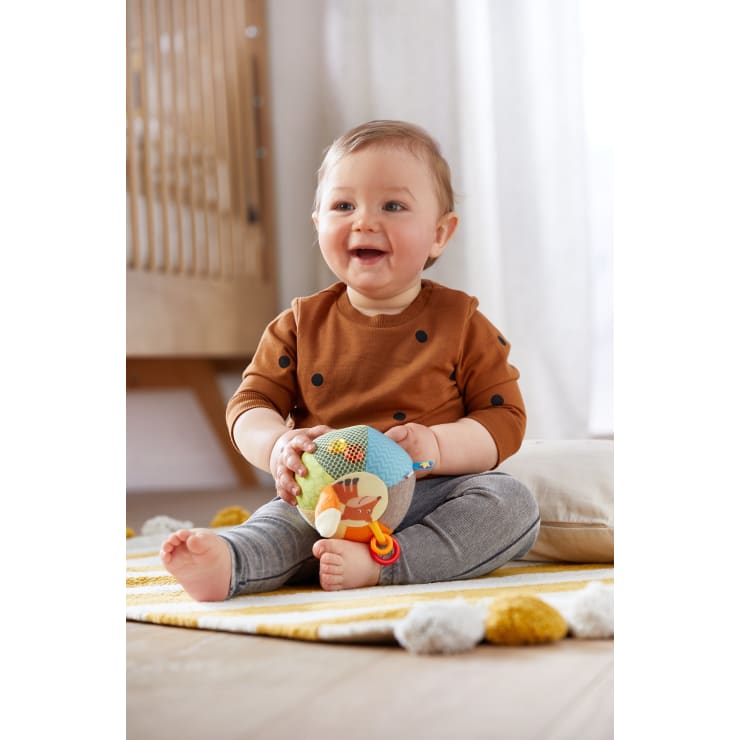 Ein Baby sitzt auf einem gelb-weiß gestreiften Teppich und hält einen bunten Stoffball in der Hand. Das Baby trägt ein braunes Hemd mit schwarzen Punkten und eine graue Hose. Im Hintergrund ist ein Kinderbett zu sehen.