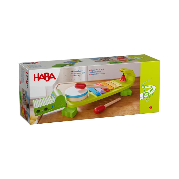 Eine rechteckige Schachtel mit einem Foto eines hölzernen Kinderspielzeugs in Form eines Krokodils, das ein Xylophon, eine Trommel und ein Waschbrett enthält. Die Marke "HABA" steht in der oberen rechten Ecke. Das Spielzeug ist für Kinder ab 2 Jahren geeignet.