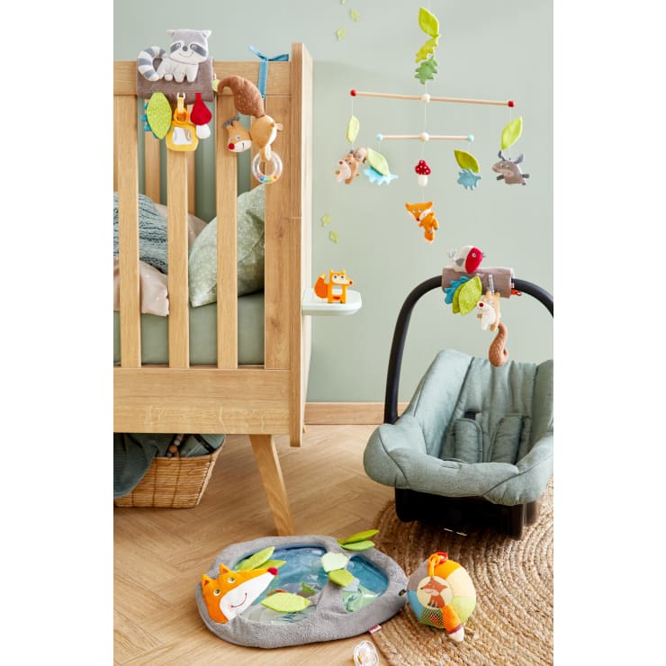 In einem Kinderzimmer mit hellgrünen Wänden und Holzfußboden stehen ein Holzbett mit Hängespielzeug in Tierform, eine Babyschale mit einem Eichhörnchen aus Plüsch, eine weiche Tierspielmatte, ein Plüschball und ein Weidenkorb.
