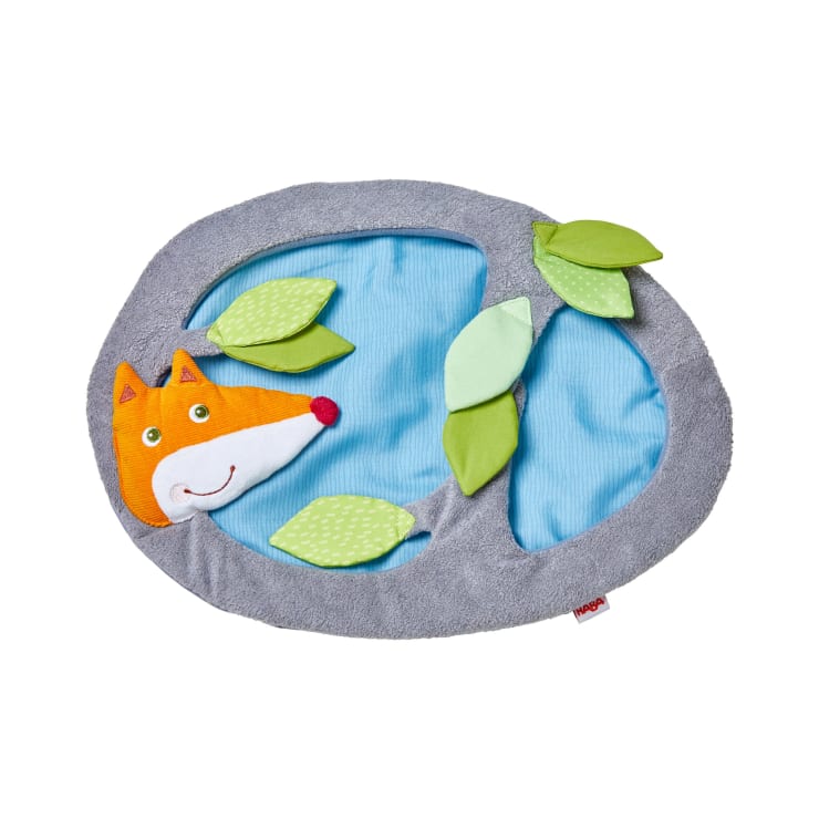 Eine runde, blau-graue Babyspielmatte aus Stoff mit einem plüschigen Fuchskopf und mehreren grünen, auf der Oberfläche angebrachten Blattformen aus Stoff. Die Matte hat eine weiche Textur und ein kleines rotes Markenetikett am Rand.
