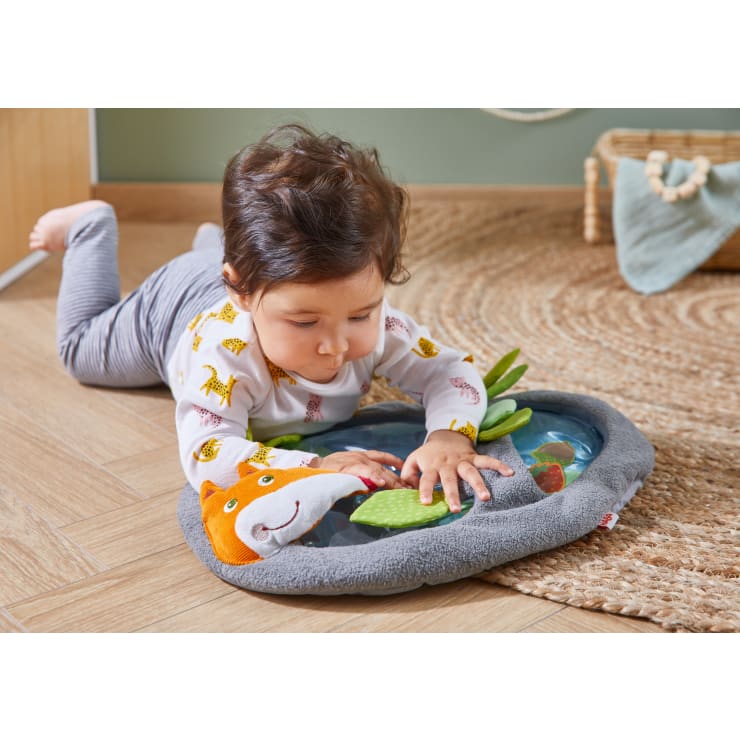 Ein Kleinkind liegt auf dem Bauch auf einem Holzboden und berührt eine sensorische Spielmatte mit Tiermotiven. Das Kind konzentriert sich auf das Spielzeug, das ein lächelndes Fuchsgesicht und verschiedene Texturen zeigt. Im Hintergrund sind ein Korb und ein Teppich zu sehen.