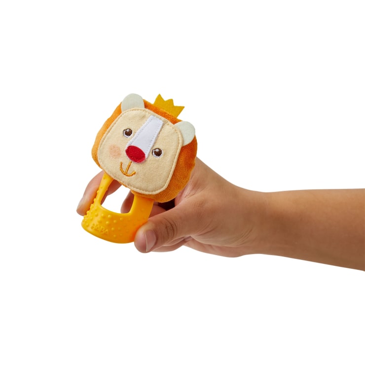 Eine Hand hält eine gelbe Babyrassel in Form eines Löwengesichts mit einer kleinen Krone auf der Oberseite, weichem Stoff am Kopf und einem strukturierten Kunststoffgriff. Der Hintergrund ist einfarbig weiß.