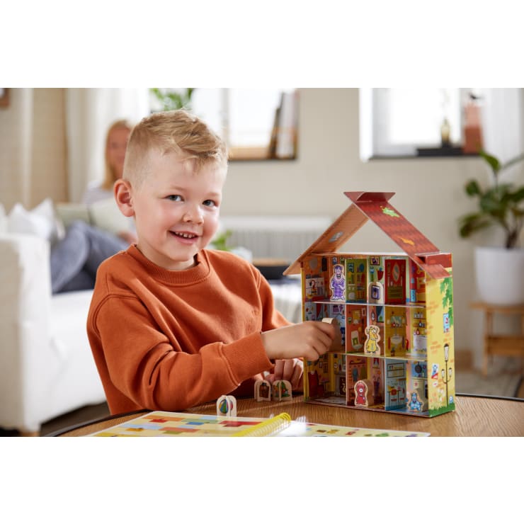 Ein kleiner Junge in einem orangefarbenen Sweatshirt spielt mit einem bunten Puppenhaus an einem Tisch. Er lächelt und stellt eine Figur in das Haus. Im Hintergrund sitzt ein Erwachsener auf einem weißen Sofa.