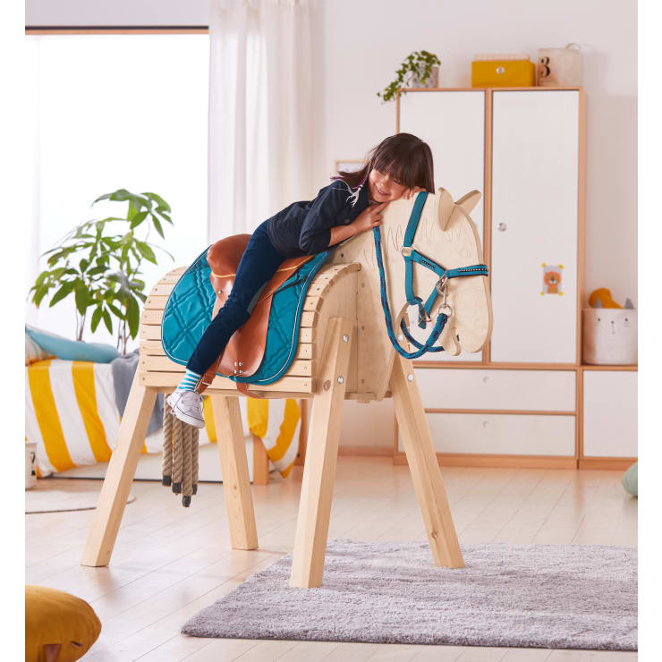 Ein Kind liegt auf einem großen hölzernen Spielzeugpferd mit einem blauen Sattel in einem hellen, aufgeräumten Zimmer, das mit Pflanzen, einem Bett und einem Teppich dekoriert ist.