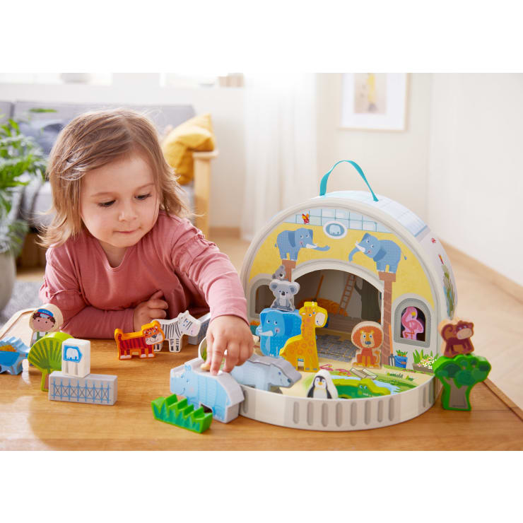 Ein kleines Kind mit hellbraunem Haar und einem rosafarbenen Hemd spielt mit Tierfiguren vor einer Spielzeugarche auf einem Holztisch in einem hellen, aufgeräumten Zimmer.