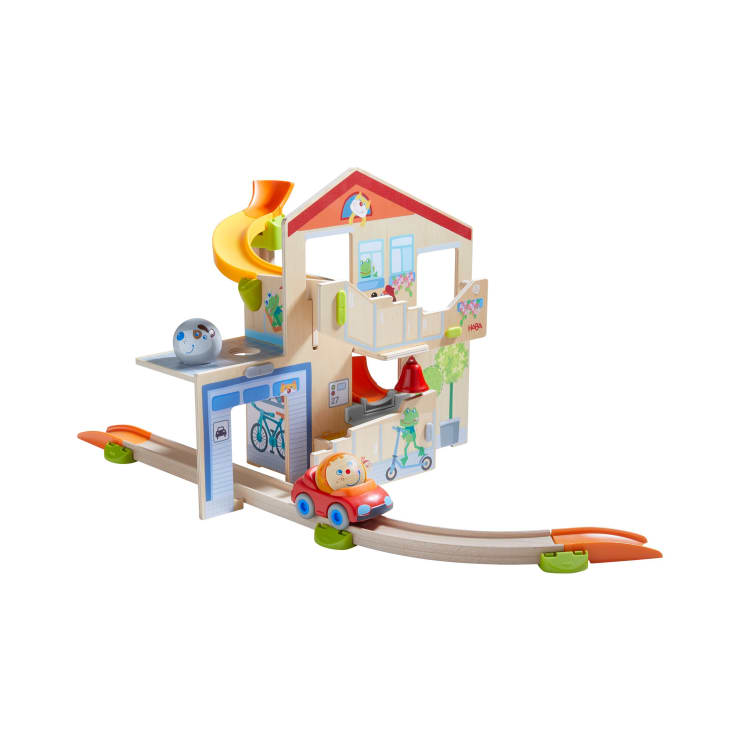 Ein Spielset aus Holz mit einem Haus mit bunten Details, einer gelben Rutsche, einer Garage und zwei Schienen mit einem orangefarbenen Spielzeugauto und einer Figur. Das Haus enthält illustrierte Elemente wie ein Fahrrad und eine Glocke.