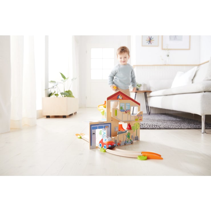 Ein kleines Kind steht in einem hellen Wohnzimmer und lächelt hinter einem hölzernen Puppenhaus mit Spielzeugautos und -schienen, das auf dem Boden vor einer Couch, Zimmerpflanzen und gerahmten Wandbildern steht.