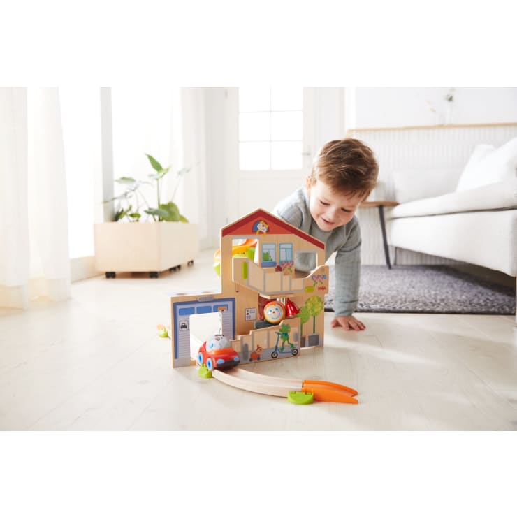 Ein kleines Kind auf dem Boden lächelt, während es mit einem bunten Holzpuppenhaus und einem Spielzeugautoset in einem hellen Wohnzimmer mit Pflanzen und einem Sofa im Hintergrund spielt.