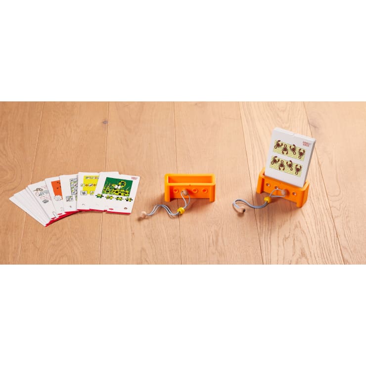 Ein Satz illustrierter Karten, ein orangefarbener Kartenhalter und ein orangefarbener Ständer mit eingelegten Karten werden auf einer Holzfläche ausgestellt. Zwei Kordeln mit gelben Perlen sind am Kartenhalter befestigt.