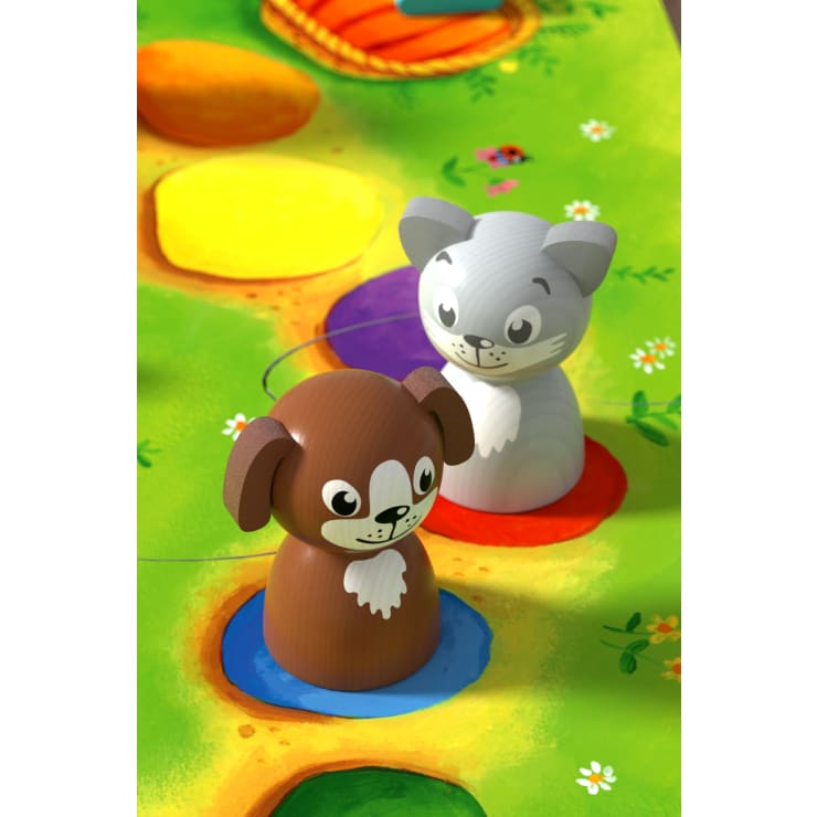 Zwei hölzerne Spielfiguren in Form eines braunen Hundes und einer grauen Katze werden auf bunten, kreisförmigen Flecken auf einem grünen, mit Blumen und einem Marienkäfer verzierten Spielbrett platziert.