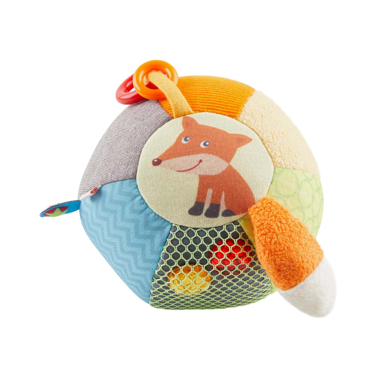 Ein weiches, mehrfarbiges Baby-Ballspielzeug mit einem lächelnden Cartoon-Fuchs, einem orangefarbenen und weißen Schwanz, einer Netztasche mit kleinen Bällen darin, einem roten Kunststoffring und verschiedenen strukturierten Stoffbahnen.
