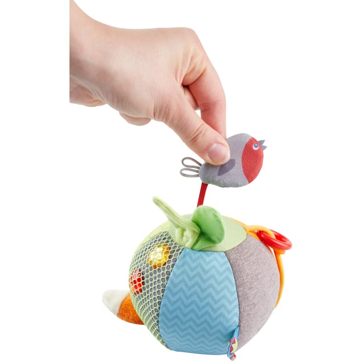 Eine Hand hält einen kleinen Stoffvogel, der an einem weichen, mehrfarbigen, strukturierten Ballspielzeug mit verschiedenen Mustern, Netzen und Stoffelementen befestigt ist.