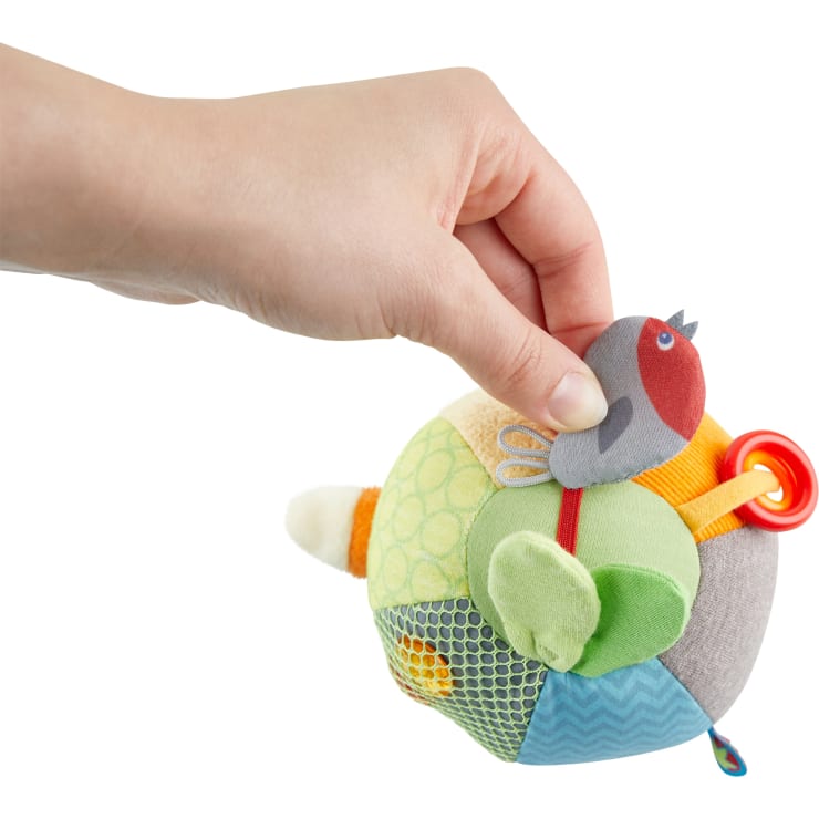 Eine Hand hält ein buntes, vogelförmiges Stofftier in Patchwork-Optik, an dessen Seite ein roter Ring befestigt ist. Das Plüschtier hat verschiedene Stoffstrukturen und einen kleinen grauen und roten Vogel auf der Oberseite.