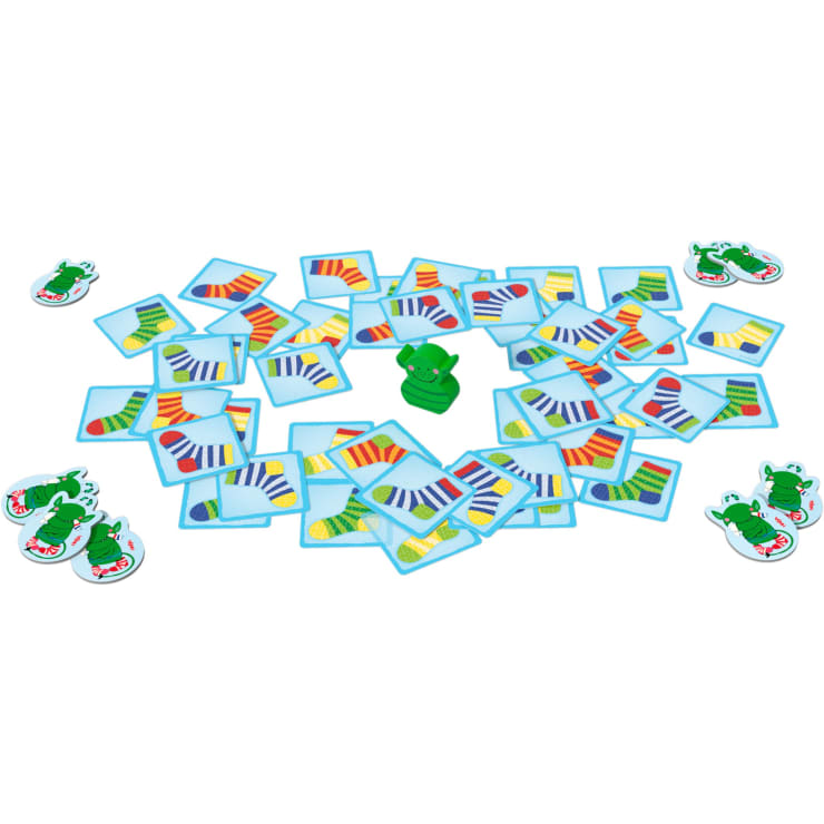 Eine Gruppe von Karten, auf denen Socken mit verschiedenen Mustern abgebildet sind, wird um eine grüne Alligatorfigur aus Plastik auf einer weißen Fläche verteilt, zusammen mit runden Spielsteinen, die grüne Schuhe zeigen.