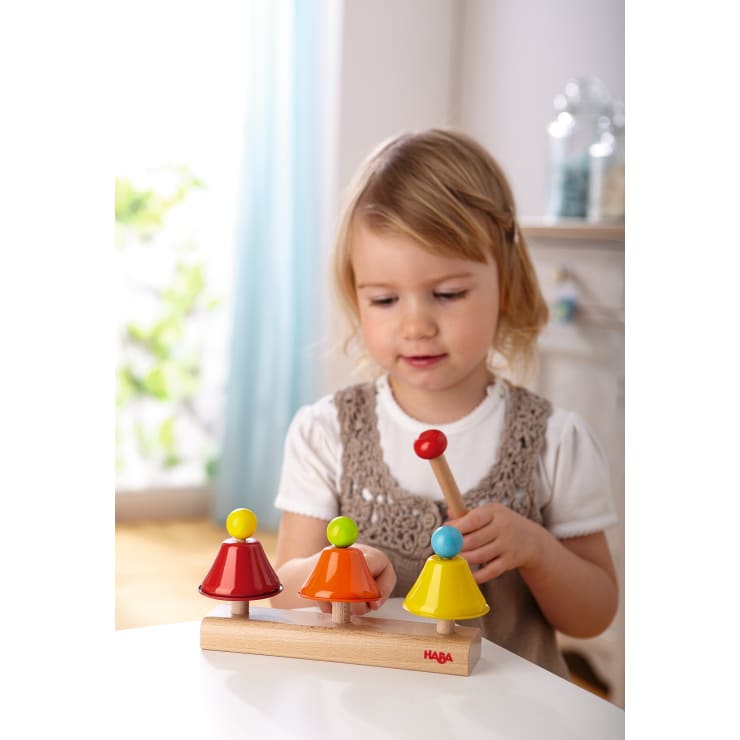 Ein kleines Kind spielt mit einem Holzschläger mit drei bunten glockenförmigen Teilen in Rot, Orange und Gelb, die jeweils mit einer Kugel versehen sind, mit einem hölzernen Musikspielzeug.
