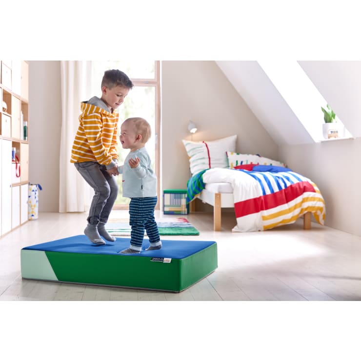 Zwei kleine Kinder, ein älteres und ein Kleinkind, stehen und hüpfen auf einer blauen und grünen Schaumstoffmatte in einem hellen, aufgeräumten Schlafzimmer mit einem Bett und buntem Bettzeug im Hintergrund.