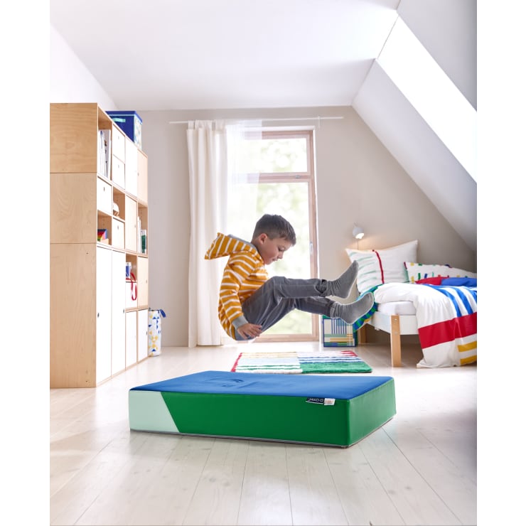 Ein kleines Kind in einem gestreiften gelben Sweatshirt und einer grauen Hose springt auf eine blau-grüne Gymnastikmatte in einem hellen Zimmer mit einem Bett, Regalen und einem Fenster, das Tageslicht hereinlässt.