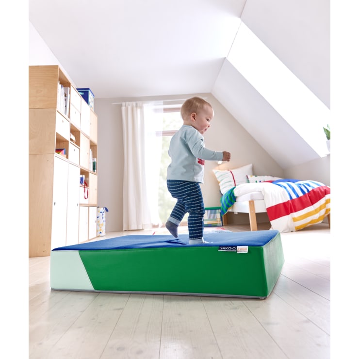 Ein Kleinkind läuft auf einem grünen und blauen Schaumstoffkeil in einem hell erleuchteten Schlafzimmer mit Holzboden, einem Bett mit gestreifter Decke und Regalen mit Büchern und Spielzeug.