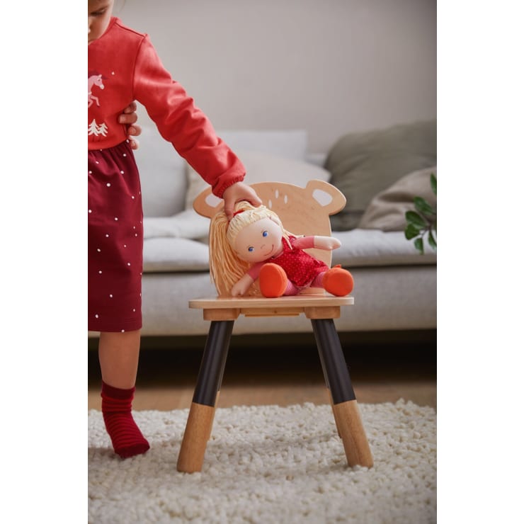 Ein Kind in einem roten Kleid und roten Socken setzt eine blonde Puppe in einem roten Outfit auf einen kleinen Holzstuhl mit tierförmigen Details. Die Szene spielt in einem Wohnzimmer mit einem weißen Teppich und einem Sofa im Hintergrund.
