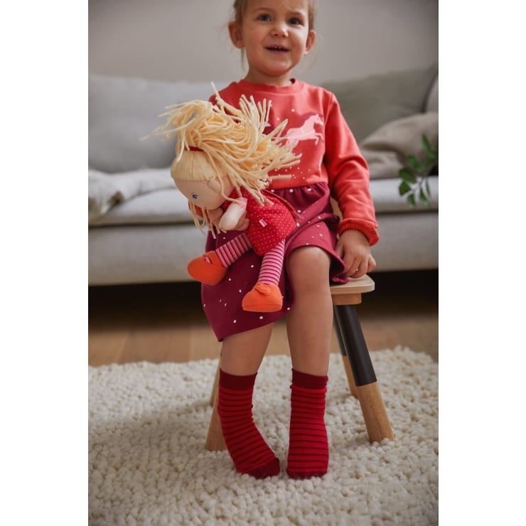 Ein kleines Kind in einem rosa-roten Kleid und roten Socken sitzt auf einem kleinen Holzschemel und hält eine Puppe mit blondem Garnhaar und rotem Outfit. Im Hintergrund sind eine beigefarbene Couch und ein Teppich zu sehen.