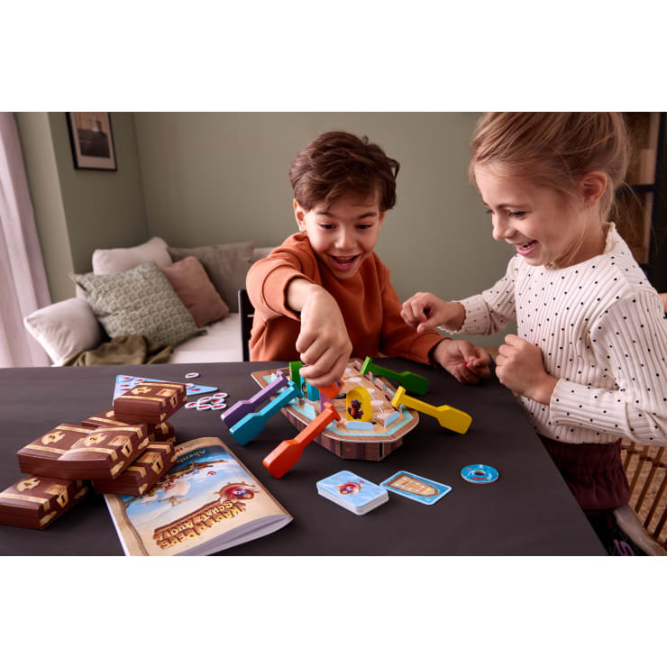 Zwei Kinder spielen ein Piratenspiel mit bunten Hebeln, Karten und Pappmünzen auf einem schwarzen Tisch. Um das Spiel herum befinden sich Stapel von Karten, Spielsteinen und Heften. Beide Kinder lächeln.