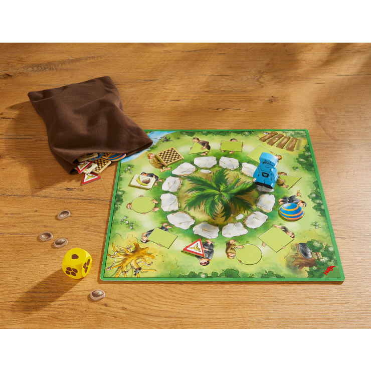 Ein Brettspiel mit einem grünen Dschungelspielbrett, einer blauen Fahrzeugspielfigur, einem gelben Würfel, runden Spielsteinen und einem braunen Stoffbeutel mit zusätzlichen Spielfiguren, alles auf einem Holztisch.