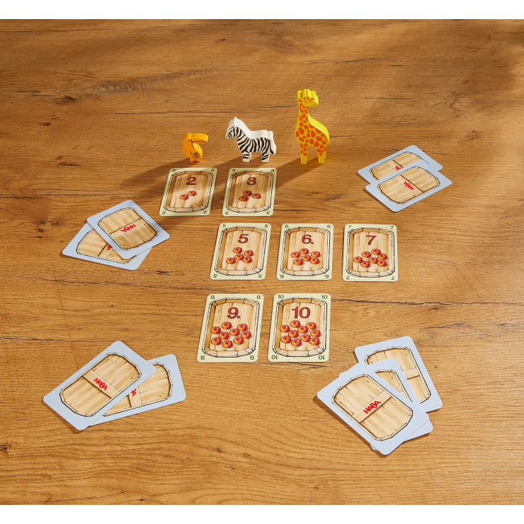 Auf einer Holzfläche liegt ein Kartenspiel mit nummerierten Karten, auf denen jeweils Äpfel abgebildet sind, und drei Tierfiguren: eine gelbe Giraffe, ein Zebra und eine orangefarbene Nummer fünf. Vier verdeckte Karten liegen in den Ecken.