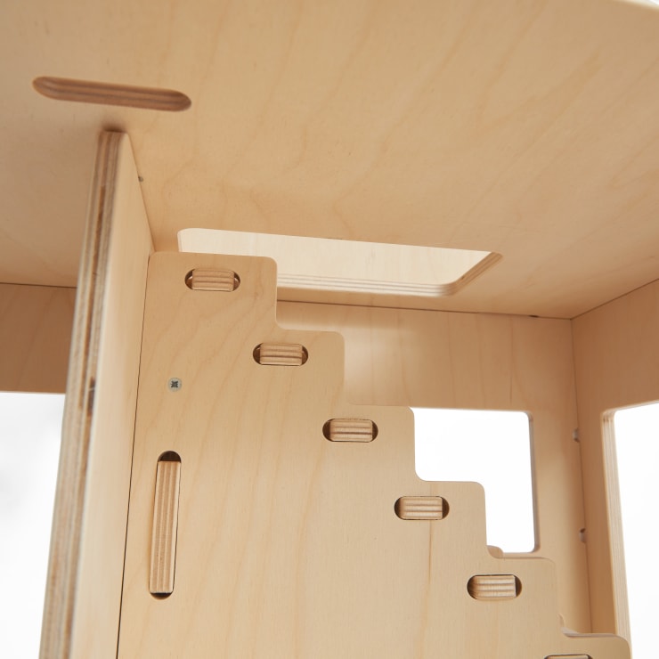 Nahaufnahme des Inneren eines hölzernen Puppenhauses, das eine Treppe mit ausgeschnittenen Schlitzen und rechteckigen Fenstern zeigt. Das helle Holz hat eine glatte Oberfläche und eine sichtbare Maserung.