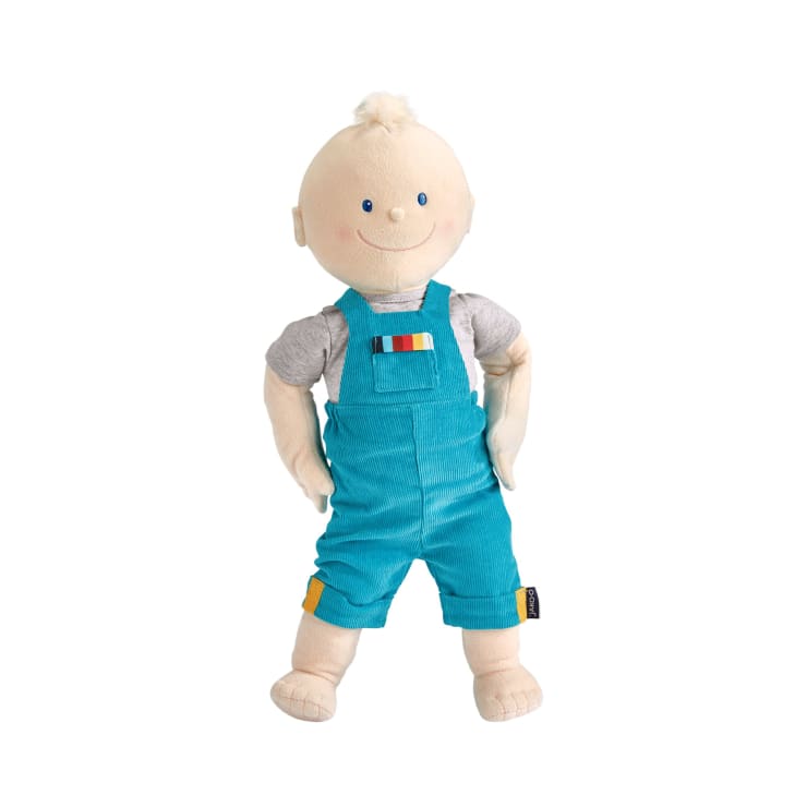 Eine Plüschpuppe mit heller Haut, blauen Augen und einem kleinen Büschel blonder Haare, die ein graues T-Shirt und eine blaue Latzhose trägt und aufrecht mit den Händen in den Hüften steht.