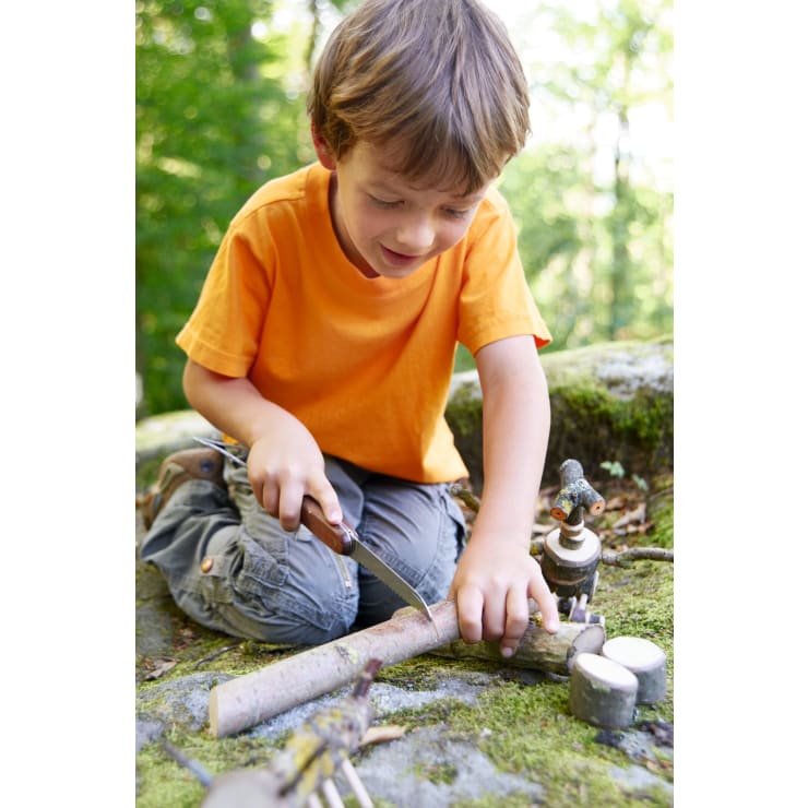 Ein kleiner Junge in einem orangefarbenen Hemd kniet auf einem Felsen im Wald und schneidet mit einem kleinen Messer einen Stock. Vor ihm auf der moosbewachsenen Oberfläche sind verschiedene Holzgegenstände und Spielzeuge angeordnet.
