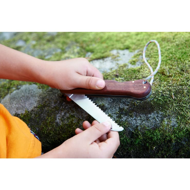 Eine Person klappt eine Taschensäge mit Holzgriff und gezacktem Sägeblatt neben einem moosbedeckten Felsen auf. Die Säge ist mit einer Handschlaufe an ihrem Griff befestigt.