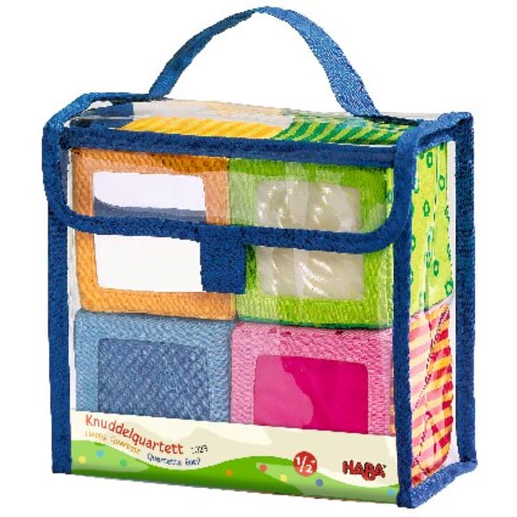 Eine transparente Tragetasche mit blauem Rand enthält vier weiche Stoffblöcke in verschiedenen Farben und Mustern. Auf dem Etikett steht "Knuddelquartett" und "Haba", was bedeutet, dass das Produkt für Kinder ab 1/2 Jahren geeignet ist.