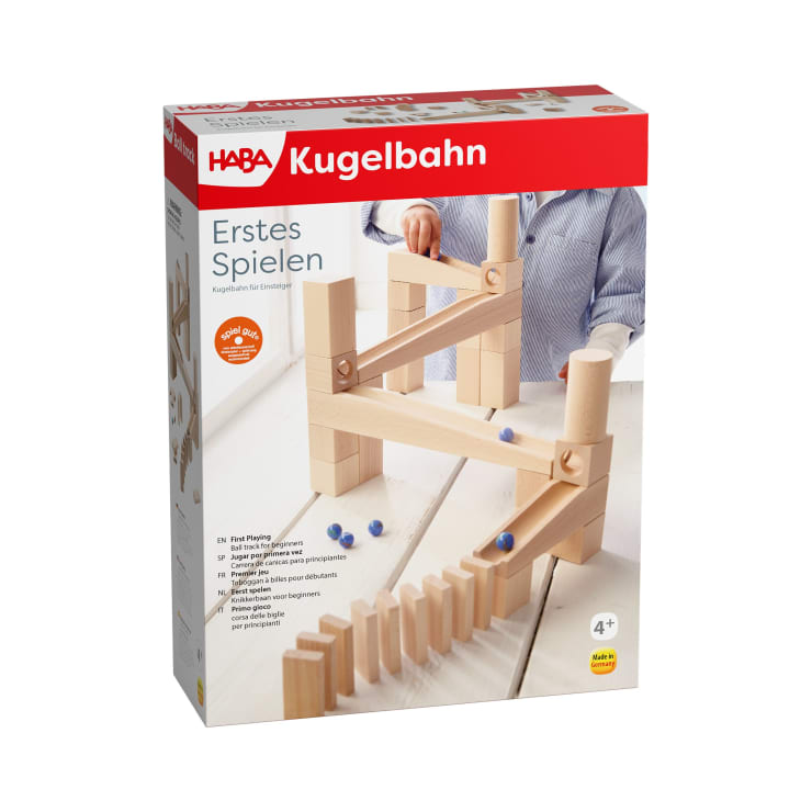 Ein Kind spielt mit einer hölzernen Kugelbahn, die auf der Vorderseite einer HABA Kugelbahn-Spielzeugkiste abgebildet ist. Auf der Schachtel sind Produktinformationen und das Branding in deutscher Sprache zu sehen sowie Bilder von Murmeln, die über Holzrampen rollen.