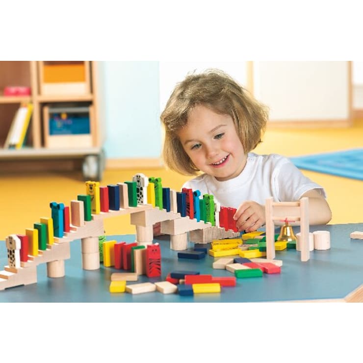 Ein kleines Kind mit hellbraunem Haar ordnet bunte Dominosteine und Holzklötze auf einem Tisch zu einem Brückenbauwerk an. Das Kind lächelt und konzentriert sich auf seine Tätigkeit. Im Hintergrund sind Regale und Spielzeug zu sehen.