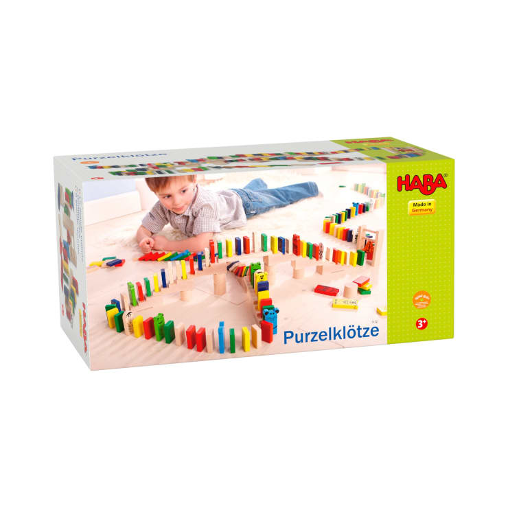 Auf einer Schachtel mit HABA Purzelklötzen ist ein Kind abgebildet, das mit bunten Dominosteinen aus Holz spielt, die in Mustern auf einem hellen Boden angeordnet sind. Die Verpackung trägt das HABA-Logo und den Hinweis, dass das Produkt für Kinder ab 3 Jahren geeignet ist.
