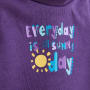 Nahaufnahme eines lilafarbenen Stoffes mit einem bunten, verspielten Text, der lautet: Everyday is a sunny day (Jeder Tag ist ein sonniger Tag) und einer Illustration einer gelben Sonne neben dem Wort day (Tag).