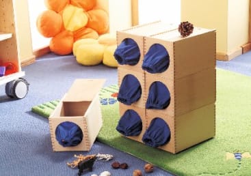 Eine Szene im Klassenzimmer zeigt eine taktile Sinnesbox mit sechs Fächern, die mit blauen Stofföffnungen abgedeckt sind, umgeben von kleinen Gegenständen auf einem grünen Teppich und gelben Sitzkissen im Hintergrund.