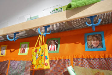 Unter einem Holzregal ist eine Reihe von blauen Haken angebracht, unter denen orangefarbene Stoffe und Kinderfotos hängen. An einem Haken hängt eine gelbe Tasche mit einem Giraffenmuster. Darunter sind Netztaschen und Künstlerbedarf zu sehen.