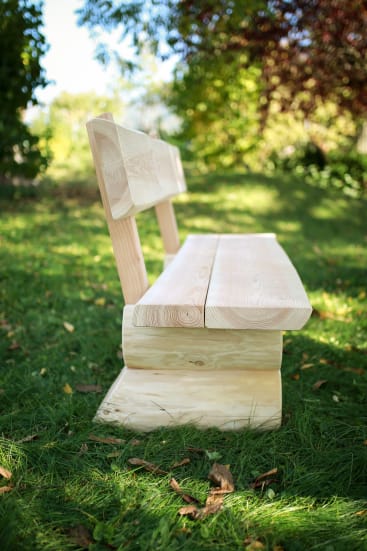 Eine helle Holzbank mit einer Rückenlehne steht auf grünem Gras in einer Außenumgebung, in der das Sonnenlicht durch die Bäume im Hintergrund fällt.