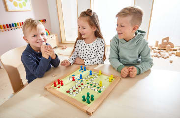 Drei kleine Kinder sitzen an einem Tisch und spielen ein Brettspiel mit bunten Figuren und Würfeln. Sie scheinen zu lächeln und sich in einem hellen Spielzimmer mit Spielzeug und Holzklötzen im Hintergrund zu beschäftigen.