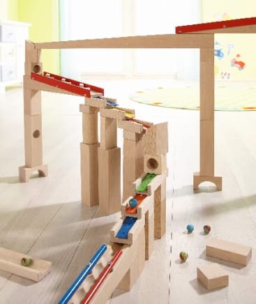 Eine hölzerne Murmelbahn ist auf einem hellen Boden aufgebaut, wobei Murmeln auf der Bahn und in der Nähe sichtbar sind. Im Hintergrund befinden sich ein Teppich, ein Fenster und eine teilweise sichtbare Kinder-Wandmalerei.
