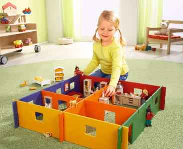 Ein kleines Kind sitzt auf einem grünen Teppich und spielt mit einem bunten, modularen Puppenhaus und kleinen Figuren. Im Hintergrund eines hellen Zimmers sind Regale mit Spielzeug und ein Teddybär zu sehen.