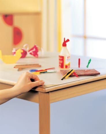 Eine Hand glättet die Kante einer weißen Tischplatte. Auf dem Tisch liegen Bastelutensilien wie Kleber, Schere, Marker und ein Universalmesser sowie ein roter Schleifblock und Schleifpapier. Der Hintergrund ist unscharf.