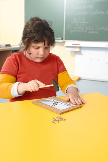 Ein Kind mit dunklen Haaren und einem rot-orangefarbenen Hemd benutzt einen kleinen Hammer, um Stecknadeln in eine Korkplatte auf einem gelben Tisch zu stecken. Im Hintergrund sind eine Kreidetafel und die Möbel des Klassenzimmers zu sehen.