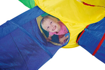 Ein kleines Kind lächelt, während es durch einen farbenfrohen Spieltunnel aus blauem, gelbem und rotem Stoff mit Netzabschnitten krabbelt.