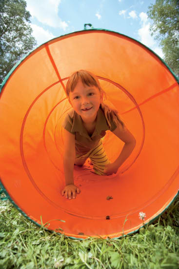 Ein kleines Kind krabbelt durch einen orangefarbenen Spieltunnel auf einer Wiese, im Hintergrund sind Bäume und der Himmel zu sehen. Das Kind lächelt und schaut in die Kamera.
