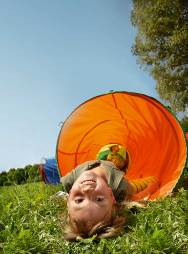 Ein Kind liegt kopfüber am Ende eines orangefarbenen Spieltunnels im Gras und lächelt in die Kamera. Ein anderes Kind ist im Hintergrund im Tunnel zu sehen. Der Himmel ist klar und blau und ein Baum steht in der Nähe.